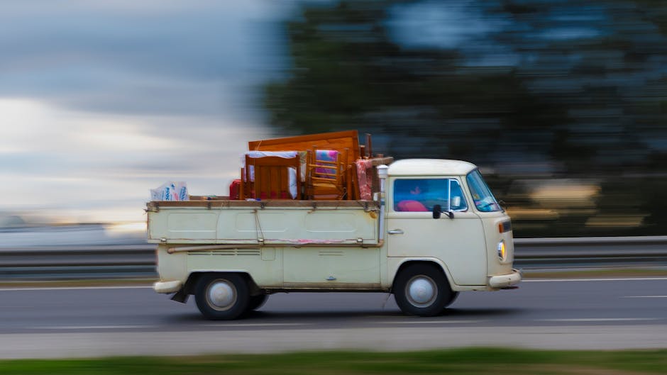 A vintage off-white van with a compact, rounded cab and small side windows is shown in motion on a road, with a blurred background indicating speed. The rear cargo area of the van is loaded with vario