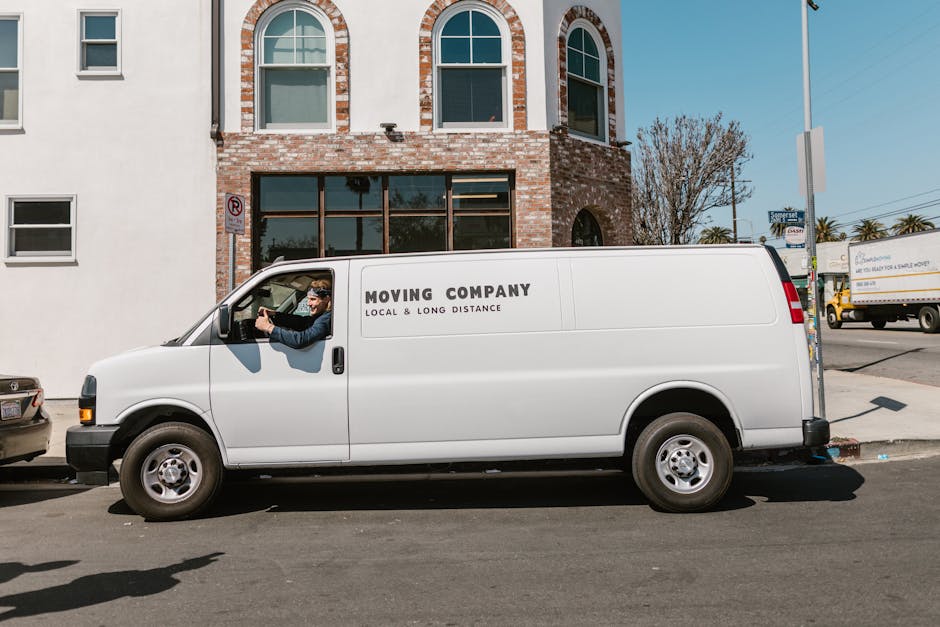 A vintage off-white van with a compact, rounded cab and small side windows is shown in motion on a road, with a blurred background indicating speed. The rear cargo area of the van is loaded with vario