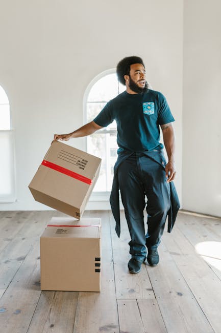 Two women standing inside a room during a home relocation process, surrounded by packed cardboard moving boxes and packing materials. The woman on the left, wearing a checked shirt and beige trousers,